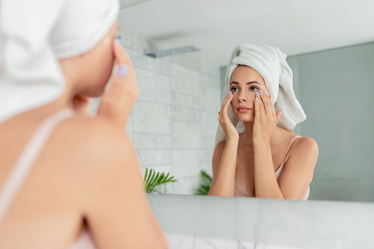 Young woman applying anti-wrinkle eye cream standing behind mirror in home bathroom